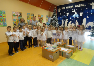 Pani dyrektor Maria Królikowska, pani Ewa, pan Zieliński i dzieci stoją wokół paczek z darami z uniesionymi kciukami.